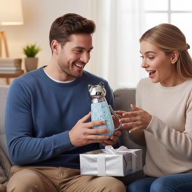 Puede incluir: Una botella de agua con forma de gato con cabeza plateada y cuerpo azul claro brillante. La botella la sostiene una persona, y una caja de regalo envuelta est&aacute; frente a ellos. La botella tiene un asa.