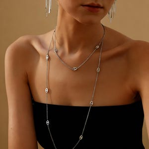 Puede incluir: Conjunto de collares plateados. El collar más corto se sitúa en la clavícula, mientras que el más largo cae sobre el pecho. Ambos collares tienen una cadena delicada con eslabones ovalados. Fondo neutro.