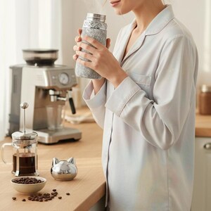 Puede incluir: Una botella de agua aislada gris y plateada con un dise&ntilde;o texturizado. La botella la sostiene una persona que lleva una camisa de pijama gris claro. Una cafetera y una prensa francesa est&aacute;n en el fondo.