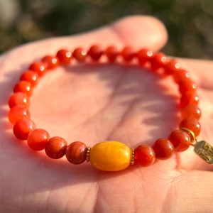 May include: A beaded bracelet featuring red and orange round beads with a central yellow oval bead. A small gold-colored tag hangs from the bracelet. The bracelet is shown against a blurred background.