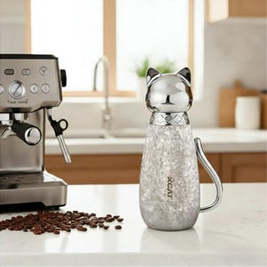 Puede incluir: Una taza de caf&eacute; con forma de gato de acero inoxidable con cabeza plateada y cuerpo moteado texturizado. La taza tiene un asa con forma de cola de gato y el texto "XCAT" est&aacute; impreso en el lateral. Una cafetera y granos de caf&eacute; est&aacute;n en el fondo.