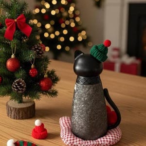 Puede incluir: Un objeto decorativo negro con forma de gato con un gorro de punto verde y un pomp&oacute;n rojo, sentado sobre un coj&iacute;n a cuadros rojos y blancos. A la izquierda hay un peque&ntilde;o &aacute;rbol de Navidad con adornos rojos y un lazo rojo.