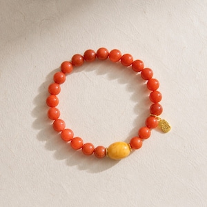 May include: A stretch bracelet composed of round, vibrant orange-red beads. A single, larger, smooth, opaque yellow oval bead is positioned near the bottom. A small, rectangular gold-toned charm with an embossed character hangs from the bracelet.