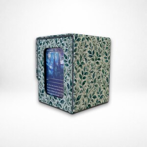 May include: A rectangular deck box featuring a green and white botanical leaf pattern across its exterior. The front side includes a clear window display showing a portion of a trading card with blue and purple stained glass artwork.