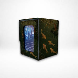 Può includere: Un contenitore rettangolare per carte collezionabili con una scena di foresta verde scuro, pesci koi arancioni e una volpe su un ramo. Una finestra centrale mostra una carta blu in stile vetrata con testo e regole di gioco visibili.