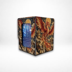 May include: A rectangular deck box for trading cards featuring a vibrant illustration of a phoenix rising over a stone city. The front panel has a clear window displaying a blue card with text. The design includes gold, orange, and red tones.
