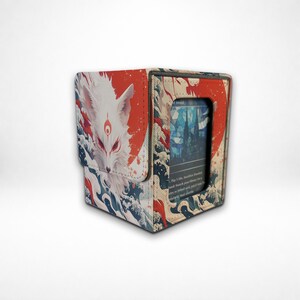 May include: A rectangular card deck box featuring a white fox illustration with red markings, set against a red sun and blue wave pattern. The front panel includes a clear window displaying a trading card with blue stained glass artwork.