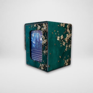 May include: A teal deck box for trading cards featuring a white cherry blossom pattern. The front has a clear rectangular window displaying a blue card with text that reads Pay 1 life, Sacrifice Flooded Strand, Search your library.