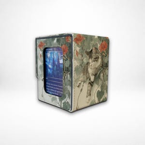 May include: A square card storage box with a decorative floral and cat design. The box features a blue card with text visible. The box is beige with red flowers and green leaves. A gray and white tabby cat is on the side of the box.