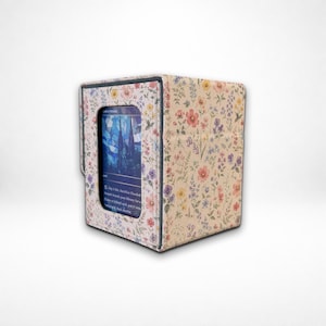 May include: A rectangular card storage box featuring a cream-colored fabric exterior with a colorful wildflower print. The front panel includes a clear rectangular window displaying a blue-toned trading card with visible printed text.