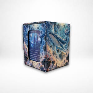 May include: A rectangular deck box featuring a blue and purple whale swimming through a sky filled with floating islands. The front panel has a cutout window displaying a trading card with text that reads, Pay 1 life, Sacrifice Flooded Strand.