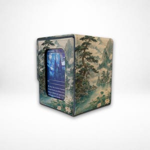 May include: A rectangular deck box featuring a printed landscape of mountains, pine trees, and a lake with lotus flowers. A central window displays a blue trading card with text that reads, Pay 1 life, Sacrifice Flooded Strand: Search your library.