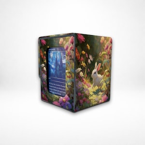 Puede incluir: Una caja de almacenamiento de tarjetas con un diseño floral y de conejo. La caja está adornada con una colorida ilustración de un conejo blanco en un campo de flores. Una tarjeta es visible en la abertura, con una imagen azul y blanca.