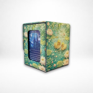 May include: A rectangular card storage box featuring a vibrant pond scene with two yellow ducklings, green lily pads, and white lotus flowers. The front panel includes a clear window displaying a blue-toned trading card with visible text.