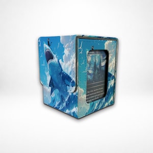 May include: A blue deck box for trading cards featuring a graphic illustration of a shark leaping from ocean waves with a small black cat on its head. The front panel has a rectangular cutout displaying a card with a stained glass cathedral design.