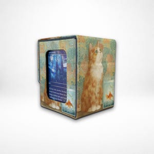 May include: A cube-shaped card storage box with a cat and goldfish design. The box features a light blue and green background with an orange tabby cat and goldfish in tanks. A card is visible in the front window.