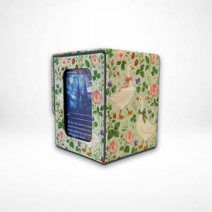 May include: A rectangular card storage box featuring a light green fabric cover with a pattern of white geese, pink flowers, red strawberries, and green leaves. A central window displays a blue-toned trading card with visible printed text.