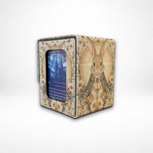 May include: A rectangular card storage box featuring a beige and cream floral pattern with a central illustration of a rabbit wearing a blue cape. A blue stained-glass style playing card is visible through the front window cutout of the box.