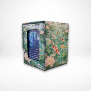 Può includere: Una scatola rettangolare per carte con un motivo stampato di pesci koi arancioni e bianchi che nuotano tra fiori di loto rosa e foglie di ninfea verdi. Una finestra centrale mostra una carta da gioco blu e viola in stile vetrata.
