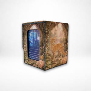 May include: A rectangular deck box featuring a printed illustration of an orange tabby cat standing in a lush, floral forest setting. The front panel includes a clear window displaying a blue-toned trading card with stained glass imagery.