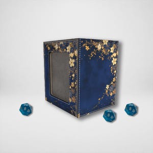 May include: A navy blue dice box with a window, decorated with gold floral patterns. The box is accompanied by three blue, twenty-sided dice. The box has white stitching around the edges.