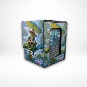 Puede incluir: Una caja para barajas rectangular con una ilustración de ranas verdes con sombreros de paja tocando guitarras bajo grandes hojas de nenúfar. La caja tiene una ventana transparente en el frente que muestra una carta con arte azul y blanco.