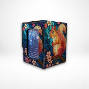 Könnte beinhalten: Eine quadratische Kartenaufbewahrungsbox mit einem Buntglasdesign, das ein rotes Eichhörnchen auf einem Ast mit rosa Blüten zeigt. Die Box hat einen blauen Hintergrund und eine Karte ist darin sichtbar.