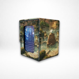 May include: A cube-shaped card storage box with a scenic illustration of a capybara relaxing in a hot spring. The box features a card with a blue and white castle design. The box is designed to hold trading cards.