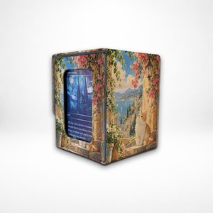 May include: A rectangular deck box featuring a printed scene of a coastal Mediterranean town with pink flowers, stone architecture, and a ginger cat looking upward. A blue trading card is visible in the front window slot of the box.