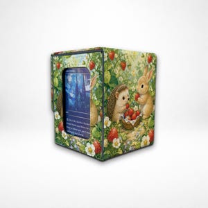 May include: A rectangular card storage box featuring a printed pattern of a hedgehog and a rabbit surrounded by strawberries and white flowers. A central window displays a blue trading card with text including "Pay 1 life, Sacrifice Flooded Strand".