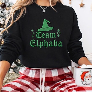May include: Black sweatshirt with a green witch hat graphic and the text "Team Elphaba". The sweatshirt is paired with red and white plaid pajama pants. A mug with a Santa Claus design is held in the hands.