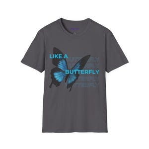 Könnte beinhalten: Ein anthrazitfarbenes T-Shirt mit einem blauen Schmetterlingsmotiv und den Worten "LIKE A BUTTERFLY" übereinander angeordnet. Der Schmetterling hat detaillierte Flügel und einen Farbverlauf in Blautönen. Das T-Shirt hat einen Rundhalsausschnitt und kurze Ärmel.