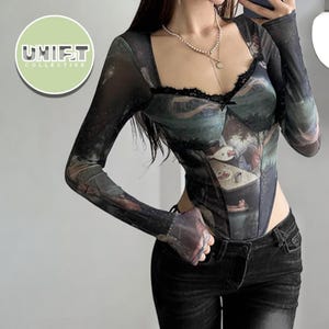 Pode incluir: Um body de manga comprida com decote quadrado e uma sobreposição transparente. O body apresenta uma estampa escura e artística e acabamento em renda preta ao longo do decote. O logotipo UNIFT COLLECTIVE está no canto superior esquerdo.