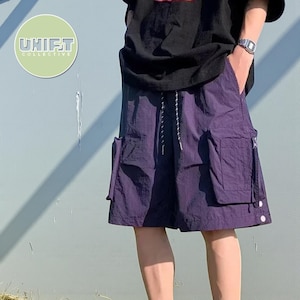 Può includere: Pantaloncini cargo viola con più tasche e vita con coulisse. I pantaloncini sono lunghi fino al ginocchio e hanno una vestibilità ampia. L'immagine presenta anche una maglietta nera e un orologio. Il logo "UNIFT COLLECTIVE" è in alto a sinistra.