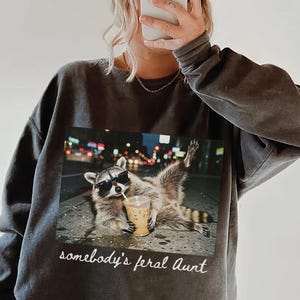 Pode incluir: Uma camisola de gola redonda cinzento-carvão com uma impressão gráfica de um guaxinim a usar óculos de sol e a segurar um café gelado numa rua à noite. Abaixo da imagem, o texto diz somebody's feral Aunt numa fonte cursiva branca.