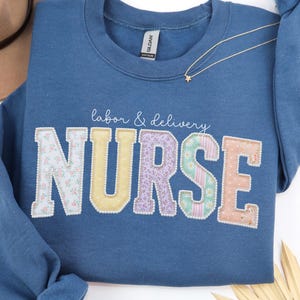 May include: A blue crewneck sweatshirt with the words "labor & delivery NURSE" in a patchwork design. The letters are in various colors and patterns, including floral and striped fabrics. A gold necklace with a star pendant is draped across the sweatshirt.
