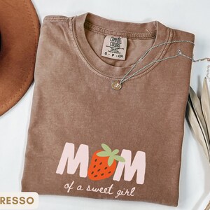 Pode incluir: Camiseta cor de caf&eacute; com o texto "MOM of a sweet girl" e um gr&aacute;fico de morango. A camisa tem gola redonda e mangas curtas. Um colar de prata com um pequeno pingente est&aacute; sobre a camisa.
