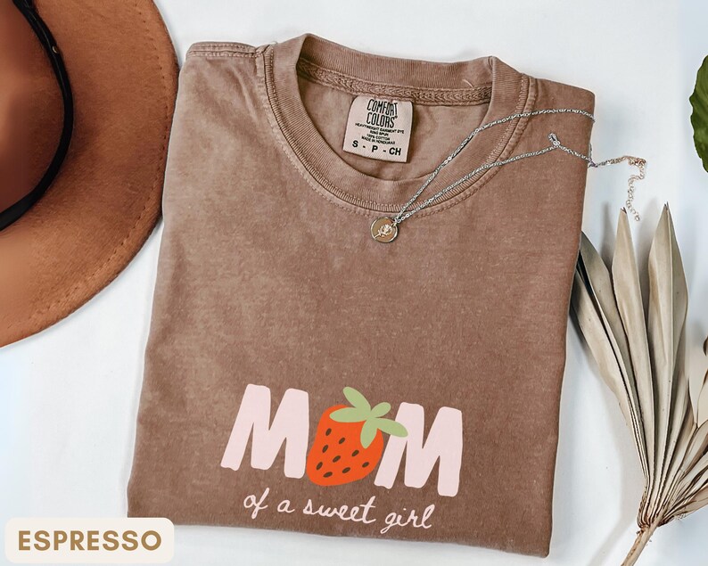 Pode incluir: Camiseta cor de caf&eacute; com o texto "MOM of a sweet girl" e um gr&aacute;fico de morango. A camisa tem gola redonda e mangas curtas. Um colar de prata com um pequeno pingente est&aacute; sobre a camisa.