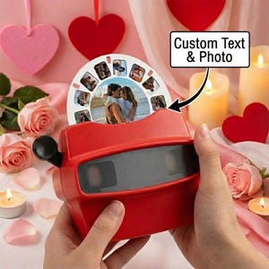 Könnte beinhalten: Ein roter Retro-Viewer mit einer benutzerdefinierten Foto-Rolle. Die Rolle zeigt mehrere Bilder eines Paares. Der Viewer wird in Händen gehalten, mit einem Hintergrund aus rosa Herzen, Rosen und Kerzen. Der Text "Custom Text & Photo" ist sichtbar.