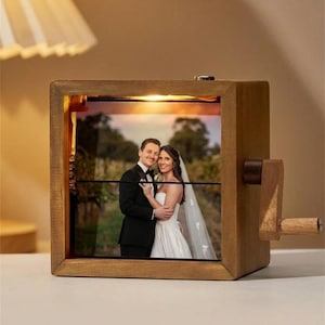May include: A wooden photo frame with a crank, displaying a wedding photo. The frame is square with a warm, natural wood finish. The photo shows a couple embracing, with a soft, warm light illuminating the image.