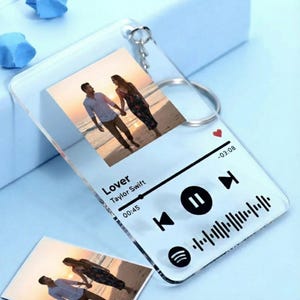 May include: A clear acrylic keychain featuring a photo of a couple on a beach. The keychain has a music player design with the song title "Lover" by Taylor Swift, a play/pause button, and a Spotify logo.