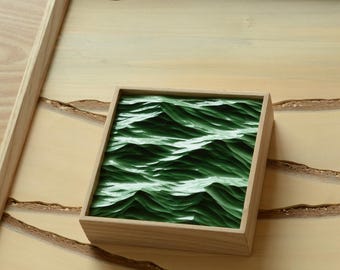 May include: A square wooden box with a carved green wave design sits on a larger wooden tray. The tray has a light wood finish with a rectangular frame and carved details. The box is centered on the tray.