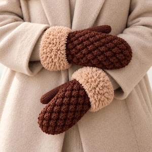 May include: A pair of dark brown knit mittens featuring a textured diamond pattern on the back of the hand and a thick, fluffy, light beige cuff. The mittens are constructed with a dense, warm-looking yarn and have a distinct thumb section.