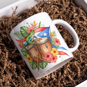 Puede incluir: Taza de cerámica blanca con una colorida ilustración de una rana verde, tambores y la bandera de Puerto Rico. El diseño incluye flores y hojas tropicales. La taza está en una caja blanca con material de embalaje marrón.
