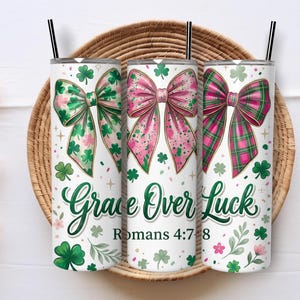 May include: Three white tumblers with black straws, each adorned with a bow design. The bows feature green, pink, and plaid patterns, complemented by shamrocks and floral accents. The text "Grace Over Luck" and Romans 4:7-8 are displayed.