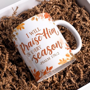 May include: White ceramic mug with the text "I WILL Praise Him IN EVERY season - PSALM 176:2" in orange script. The mug is decorated with orange and yellow floral accents and sits in a white box filled with brown crinkle paper.