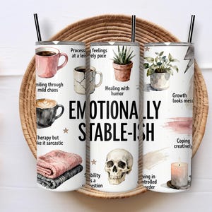Può includere: Tre bicchieri isolanti bianchi con cannucce nere, con la scritta "Emotionally Stable-ish" e illustrazioni ad acquerello di tazze di caffè, piante, un teschio e una candela. I bicchieri sono appoggiati su un vassoio intrecciato.