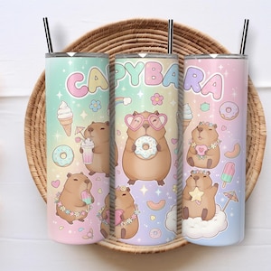 Op de afbeelding: Drie pastelkleurige geïsoleerde bekers met zwarte rietjes, elk met een capybaradesign. Het woord "CAPYBARA" staat bovenaan. De capibara's worden afgebeeld met ijs, donuts en andere zomerse lekkernijen.