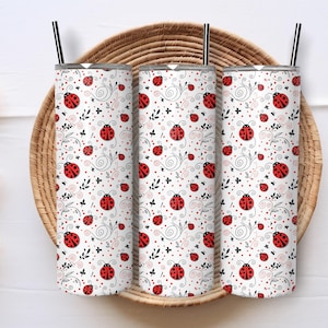May include: Three white insulated tumblers with black straws, featuring a repeating pattern of red ladybirds, black swirls, and small pink dots on a white background. The tumblers are displayed on a woven basket.