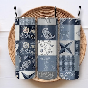 May include: Three stainless steel tumblers with black straws, featuring a patchwork design in shades of blue, white, and gray. Each tumbler has a different pattern, including floral, paisley, and deer motifs. The tumblers are displayed on a woven tray.